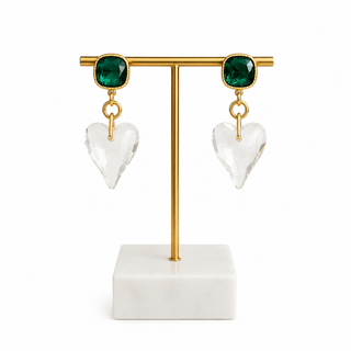 A pair of gold drop earrings with square green gemstones and clear faceted heart-shaped dangling pendants, displayed on a gold T-bar jewelry stand with a white marble base against a white background.