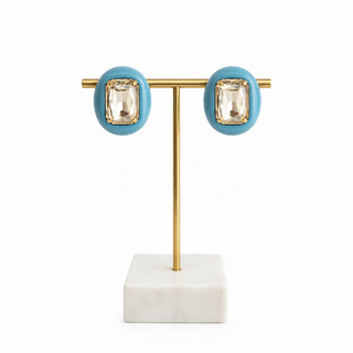 A pair of oval stud earrings with turquoise blue bezel settings and clear rectangular marquise-cut crystals, displayed on a gold T-stand with a white marble base against a white background.