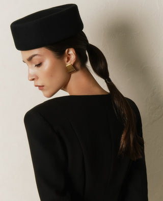 Woman wearing a black hat and black outfit with gold earrings against a neutral background