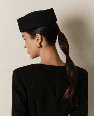 Woman wearing a black hat and black outfit with gold earrings against a neutral background
