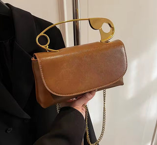 Brown handbag with gold handle and chain detail held by a person wearing a black coat.