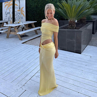 Woman in a yellow off-shoulder dress standing on a wooden deck with outdoor furniture and plants in the background.