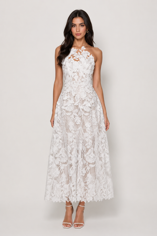 A woman modeling a sleeveless white lace maxi dress with a fitted bodice, floral lace overlay, and sheer mesh skirt, paired with clear high-heeled sandals against a light gray studio background.