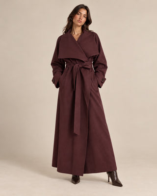 A woman modeling a long, dark plum-brown wrap coat with a wide collar, long sleeves, and a matching waist tie, paired with dark pointed-toe heeled boots against a plain beige studio background.