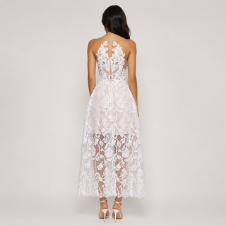 A woman viewed from the back wearing a white lace dress with floral lace applique and a cowl neck, paired with nude high-heeled sandals against a light gray studio background.