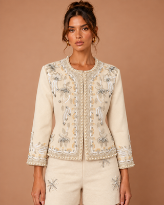 A woman modeling a cream-colored embellished jacket with intricate silver and beige embroidery, paired with matching cream shorts, against a solid warm brown studio background.