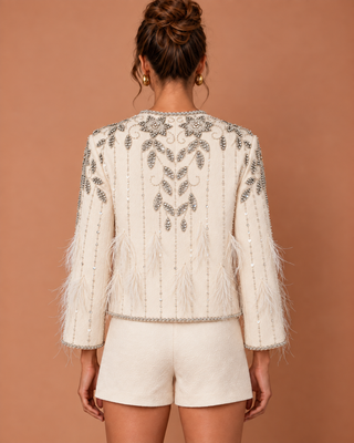 A woman shown from the back wearing a cream-colored embellished jacket with feathered sleeves and silver beadwork, paired with matching cream shorts, against a solid warm brown background.