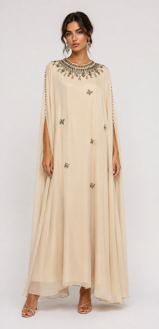 A woman modeling a floor-length beige caftan dress with a wide collar, draped sleeves, and scattered metallic star embellishments, photographed against a plain light gray studio background.