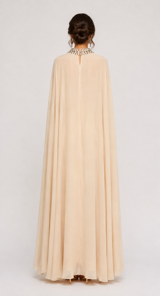 A woman viewed from the back wearing a floor-length flowing cape-style dress in a light warm cream or champagne tone, with a jeweled neckline detail, against a plain light gray studio background.