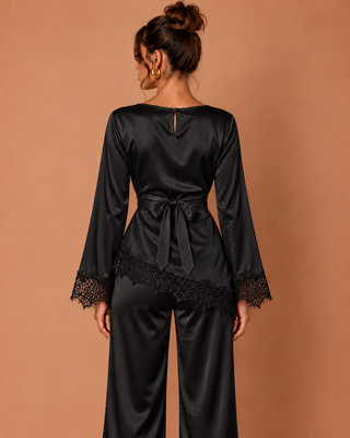 A woman shown from the back wearing a black satin jumpsuit with long sleeves, lace cuffs, a tie waist, and wide-leg pants, posed against a solid warm tan background.