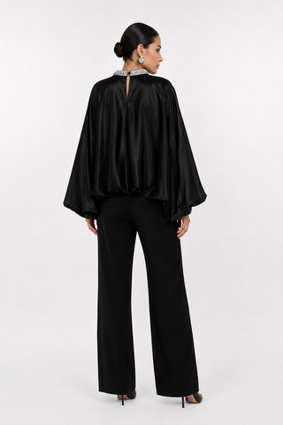 A woman shown from the back wearing a black satin cape-style top with a beaded collar and black wide-leg trousers, photographed against a light gray studio background.