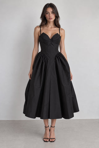 A woman modeling a black midi dress with a sweetheart neckline, structured corset-style bodice, and full gathered skirt, standing against a plain light gray studio background.