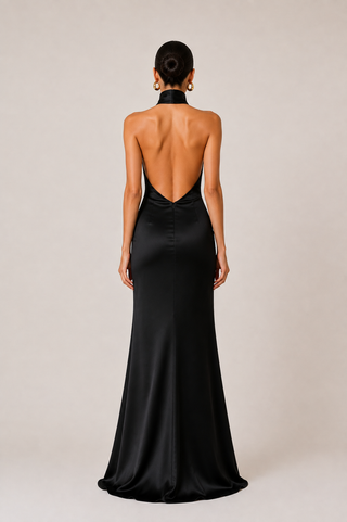 A woman viewed from the back wearing a floor-length black satin gown with a high neck and a dramatic open back, standing against a plain light beige studio background.