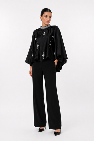 A model stands against a light gray studio background wearing a black satin-like top with wide draped sleeves, silver embroidered motifs, a beaded collar, and black wide-leg trousers.