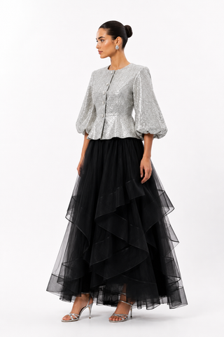 A woman models a two-piece formal outfit consisting of a silver sequined cropped jacket with puff sleeves and a long black tiered tulle skirt, set against a plain white studio background.
