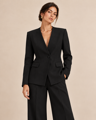 A woman modeling a black tailored blazer with structured shoulders and a single front button, paired with matching black wide-leg trousers, against a plain warm beige studio background.