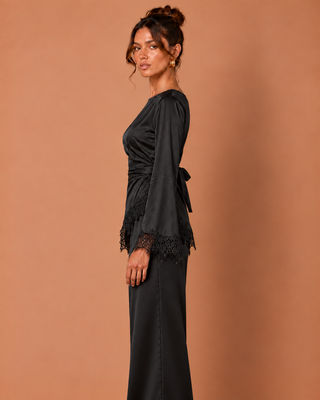 A woman modeling a black long-sleeve wrap-style dress with lace trim at the cuffs and a side tie, posed against a solid warm peach studio background.