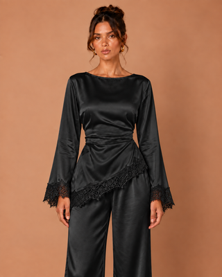 A woman modeling a black satin long-sleeve jumpsuit with lace trim on the cuffs and hem, set against a solid warm brown studio background.