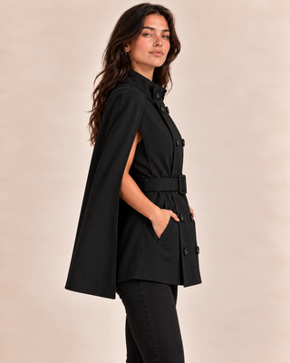 A woman modeling a black cape-style belted coat with a high collar and front buttons, shown in side profile against a plain beige studio background.