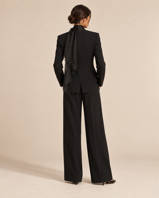 A model viewed from the back wearing a black tailored suit with wide-leg trousers and a draped scarf detail at the back of the blazer, set against a plain beige studio background.