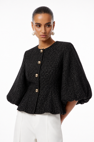 A model wearing a black textured tweed jacket with dramatic balloon sleeves and gold buttons, paired with white trousers, against a plain white background.