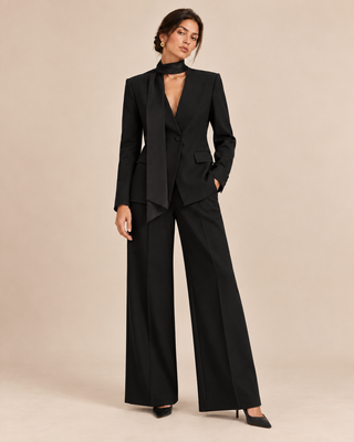 A model wearing a black tailored suit with wide-leg trousers, a structured blazer, and a matching high-neck choker, posed against a solid beige studio background.