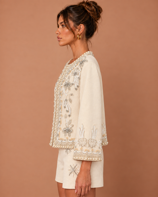 A woman shown in side profile wearing a cream-colored embroidered jacket with wide bell sleeves and matching shorts against a solid warm brown background.