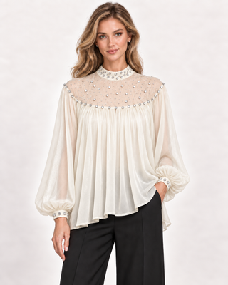 A woman modeling a cream-colored long-sleeve blouse with a heavily embellished beaded yoke and matching beaded cuffs, paired with black pants, against a plain light background.