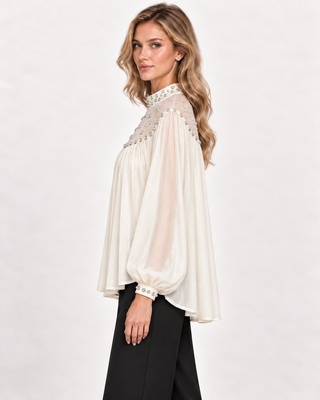 A woman modeling a cream-colored long-sleeve blouse with intricate silver beaded detailing on the shoulders and cuffs, paired with black pants, against a plain light gray studio background.