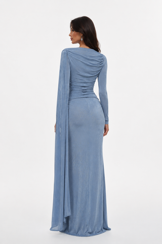 A woman shown from the back wearing a floor-length draped gown in a dusty light blue fabric, photographed against a clean white studio background.
