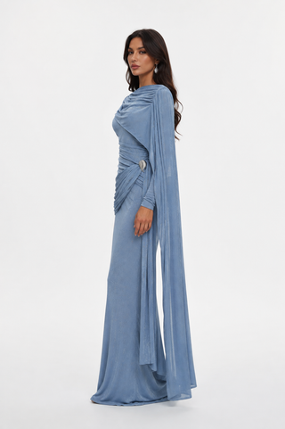 A woman modeling a floor-length light blue draped gown with long sleeves and a flowing cape-like overlay, photographed against a plain light gray studio background.
