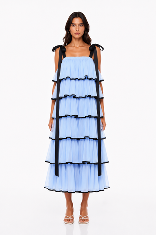 A model wearing a light blue tiered tulle dress with black ribbon shoulder straps and black ruffle trim, paired with white strappy heels, against a plain white studio background.