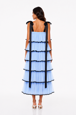A model viewed from the back wearing a light blue tiered tulle dress with black ribbon shoulder ties, set against a plain white studio background.
