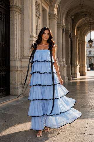 A woman stands in a stone archway corridor wearing a light blue tiered maxi dress with black ribbon accents and shoulder straps.