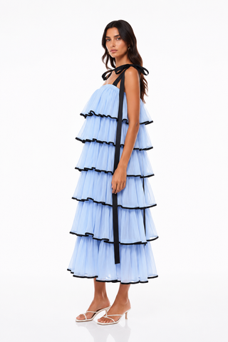 A model wearing a light blue tiered tulle maxi dress with black ribbon tie details at the shoulders and along the ruffle edges, paired with white heeled sandals, against a plain white studio background.