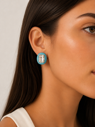 A close-up of a woman's ear wearing a turquoise blue oval stud earring with a clear rectangular baguette gemstone in the center, set against a neutral light gray background.