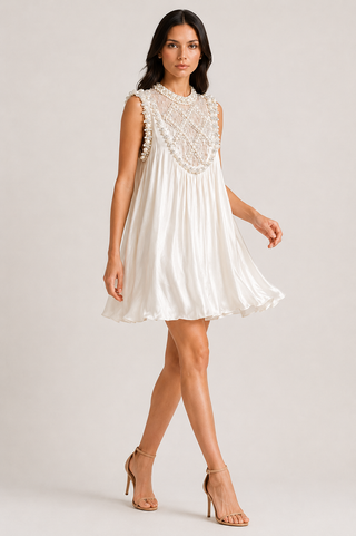 A woman modeling a sleeveless white mini dress with a pleated skirt and a high neckline decorated with intricate lace and beadwork, paired with beige high-heeled sandals against a light gray studio background.