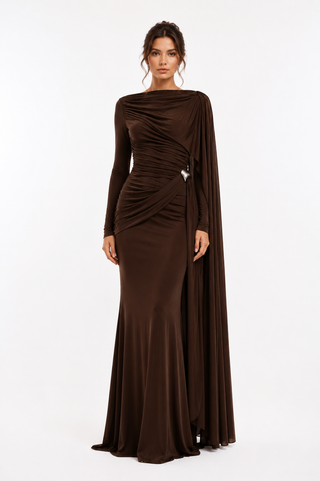 A woman modeling a floor-length dark brown gown with long sleeves, an asymmetrical draped neckline, ruched detailing at the waist, and a flowing cape-like drape over one shoulder, photographed against a clean white studio background.
