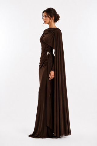 A woman modeling a floor-length dark brown evening gown with draped fabric and a matching cape-like overlay, shown in side profile against a clean white studio background.