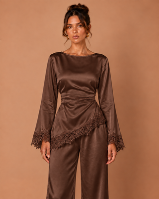 A woman modeling a brown satin long-sleeve top and matching wide-leg pants against a solid warm brown studio background.