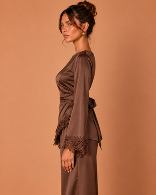 A woman in side profile wearing a brown satin long-sleeve wrap-style dress with lace trim at the cuffs, photographed against a warm tan studio background.