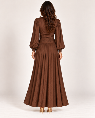 A woman viewed from the back wearing a long brown pleated maxi dress with voluminous long sleeves and a gathered waist, paired with tan heels, against a plain light background.