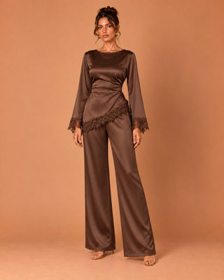 A woman models a two-piece satin jumpsuit in a warm brown shade, featuring a long-sleeve top with lace trim and matching wide-leg pants, set against a solid warm brown studio backdrop.