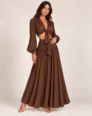 A woman modeling a brown two-piece set consisting of a long-sleeve cropped top with a twisted front and a matching pleated maxi skirt, photographed against a plain light beige studio background.