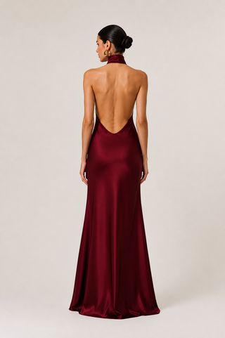 A woman viewed from the back wearing a floor-length satin gown in deep burgundy with a high neck and an open back, standing against a plain light beige studio background.