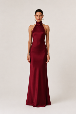 A woman modeling a floor-length, sleeveless halter-neck gown in deep burgundy satin against a plain light beige studio background.