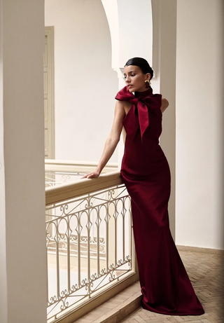 A woman in a floor-length deep burgundy gown with a large bow at the neckline stands on an indoor balcony with a white railing, looking off-camera.