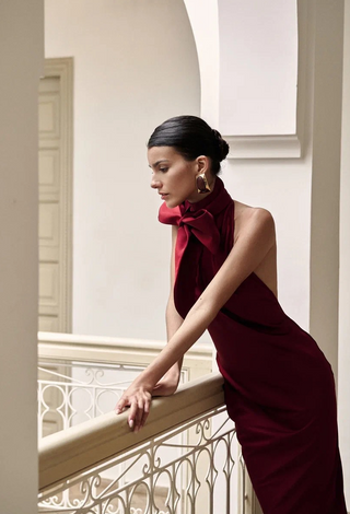 A woman in a sleeveless deep burgundy dress with a large bow at the neck leans on an ornate cream railing in a bright interior setting.