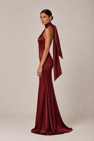 A woman modeling a floor-length satin gown in deep burgundy. The dress features a high halter-style neckline with a dramatic draped fabric detail over the shoulder and back, a fitted silhouette, and a slight train, photographed against a plain light beige studio background.