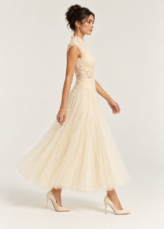 A woman modeling a sleeveless cream tulle midi dress with a lace bodice, shown from a side profile against a light gray studio background.